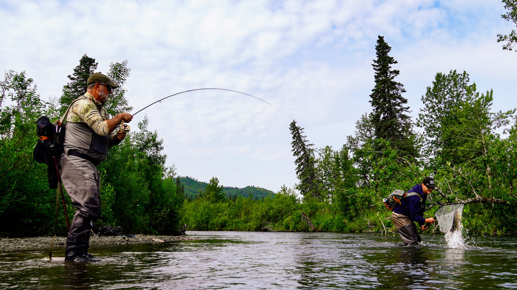 Two fly fisherman wading in a river in Bristol Bay Alaska, with one casting a fishing rod and the other netting fish, surrounded by green trees and hills under a cloudy sky.