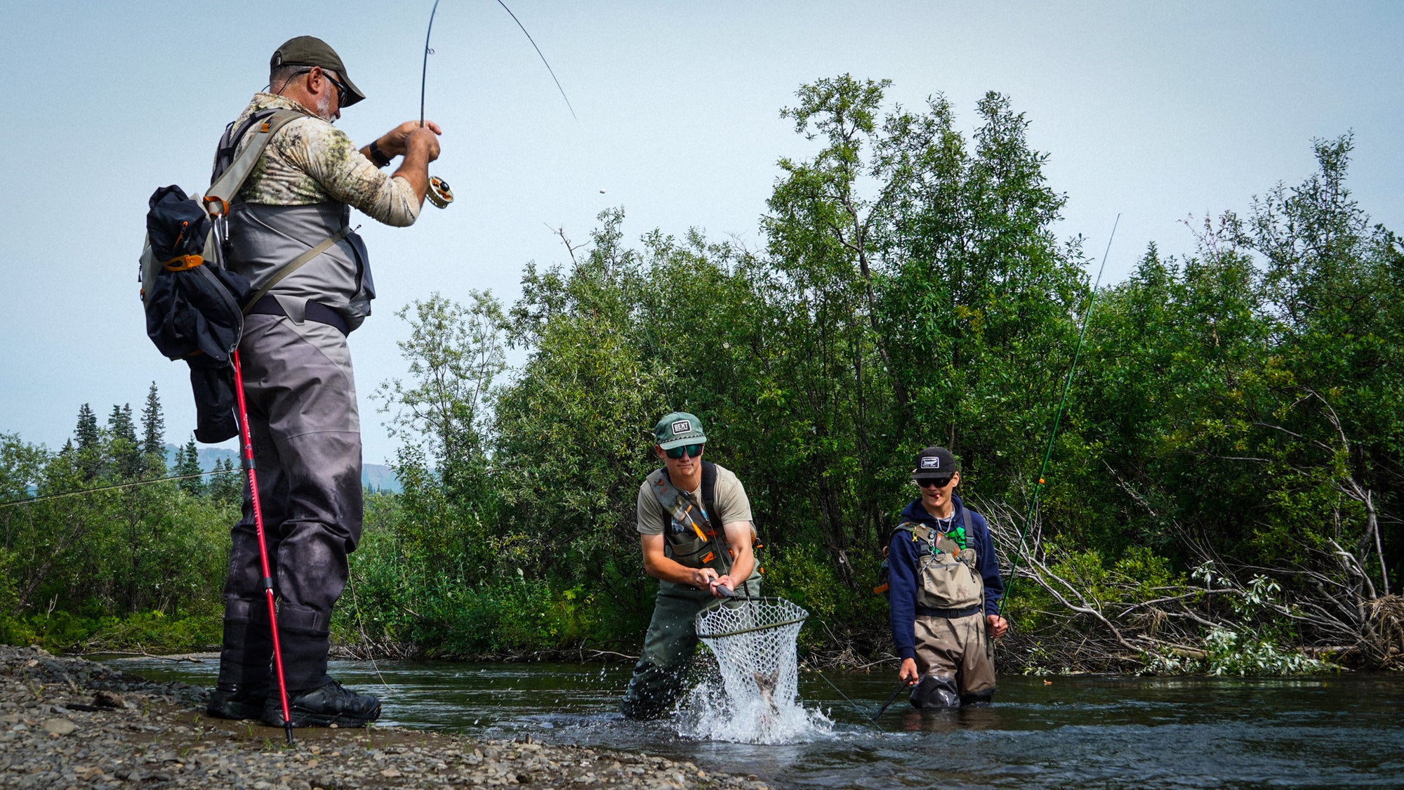Three men wading in a river in Alaska, Bristol Bay during a fly-fishing trip, with one casting a rod, one netting fish in a splash, and another standing nearby, set against a backdrop of green trees and a pale sky.