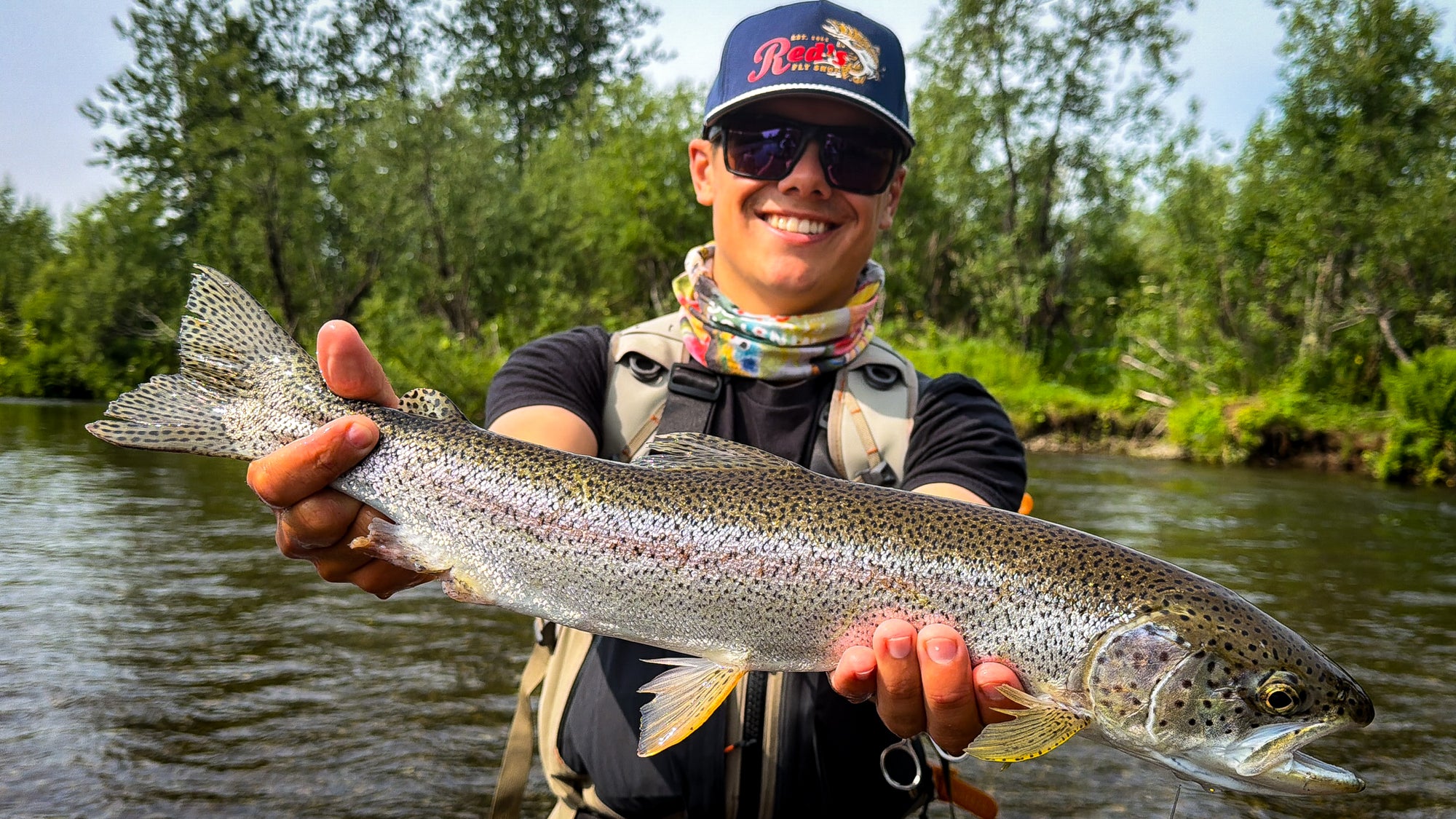 A smiling man wearing a blue Red's Fly Shop cap, sunglasses, and a neck gaiter holds a large rainbow trout over a river with trees in the background on an Alaskan River. 